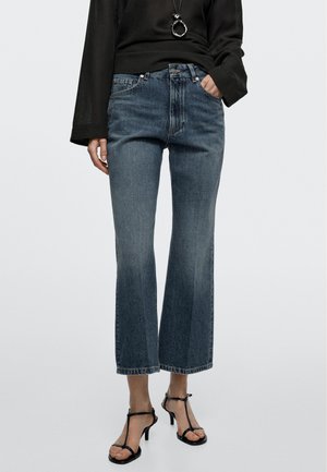 Person iført høj talje, cropped flare jeans i mørk vask, sort langærmet skjorte, sølvsmykke og sorte hælede sandaler.