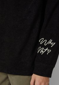 Czarna koszula z długim rękawem wykonana z miękkiego materiału, na lewym rękawie haftowane białe napisy „Why Not?”. Dopasowane mankiety.