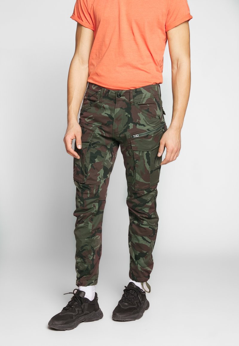 Spodnie cargo w kamuflażu w kolorach zielonym i brązowym, z wieloma kieszeniami i detalami na zamki. Połączone z koralową koszulą z krótkim rękawem i czarnymi sneakersami.