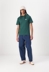 Πράσινο βαμβακερό μπλουζάκι με κοντά μανίκια, στρογγυλή λαιμόκοψη και μικρό λογότυπο. Συνδυάζεται με φαρδιά μπλε τζιν παντελόνια και λευκά παπούτσια με κόκκινες λεπτομέρειες.