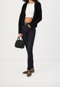 Frau trägt eine schwarze Strickjacke, ein weißes Crop-Top, dunkle Skinny-Jeans, Leopardenmuster-Ballerinas und hält eine schwarze, texturierte Handtasche.