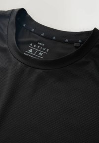 Zbliżenie na czarny, teksturowany dekolt sportowej koszulki, na którym widać metkę z rozmiarem M oraz marką "Next Active".