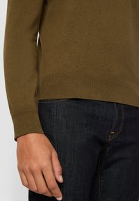 Maglione a maglia verde oliva con orlo e polsini a coste, abbinato a jeans di denim scuro. Il tessuto appare morbido e testurizzato.