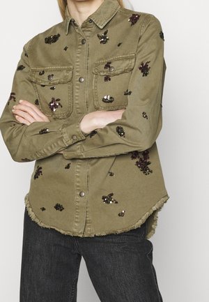 Oliwkowa koszula zapinana na guziki z postrzępionym dołem, ozdobiona rozproszonymi ciemnymi cekinowymi wzorami kwiatowymi, noszona z ciemnymi jeansami i skrzyżowanymi ramionami.