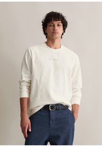 Langärmliges cremefarbenes Hemd mit Rundhalsausschnitt und subtilen Logo, kombiniert mit dunkeldblauen Jeans und einem schwarzen Gürtel. Glatte Baumwolltextur.