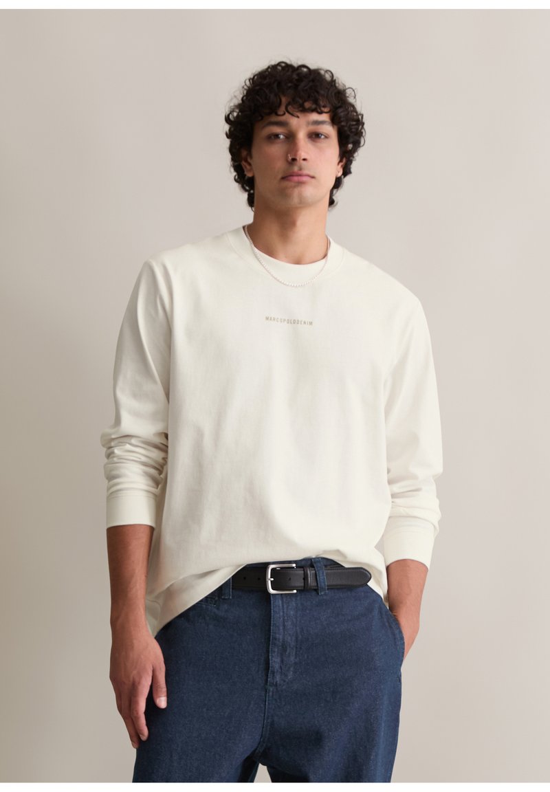 Langärmliges cremefarbenes Hemd mit Rundhalsausschnitt und subtilen Logo, kombiniert mit dunkeldblauen Jeans und einem schwarzen Gürtel. Glatte Baumwolltextur.