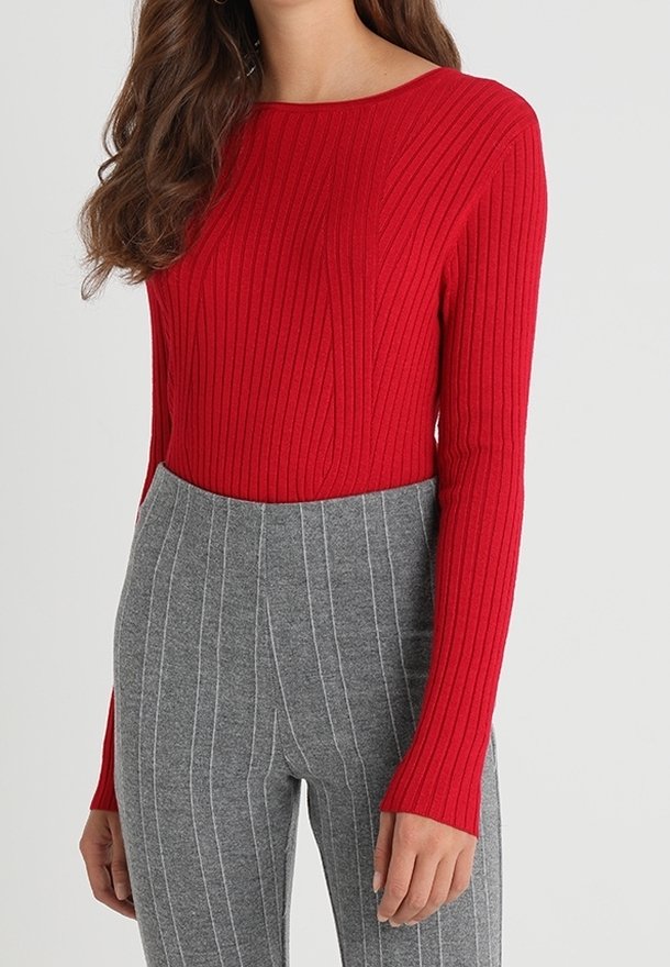 Donna che indossa un top a maniche lunghe rosso attillato e pantaloni grigi a righe alte in vita su uno sfondo chiaro e semplice.