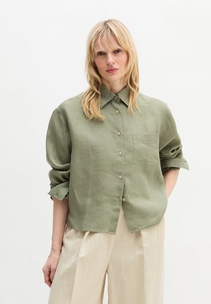 Femme blonde portant une chemise boutonnée ample de couleur olive avec les manches retroussées et un pantalon large crème, debout devant un fond uni.