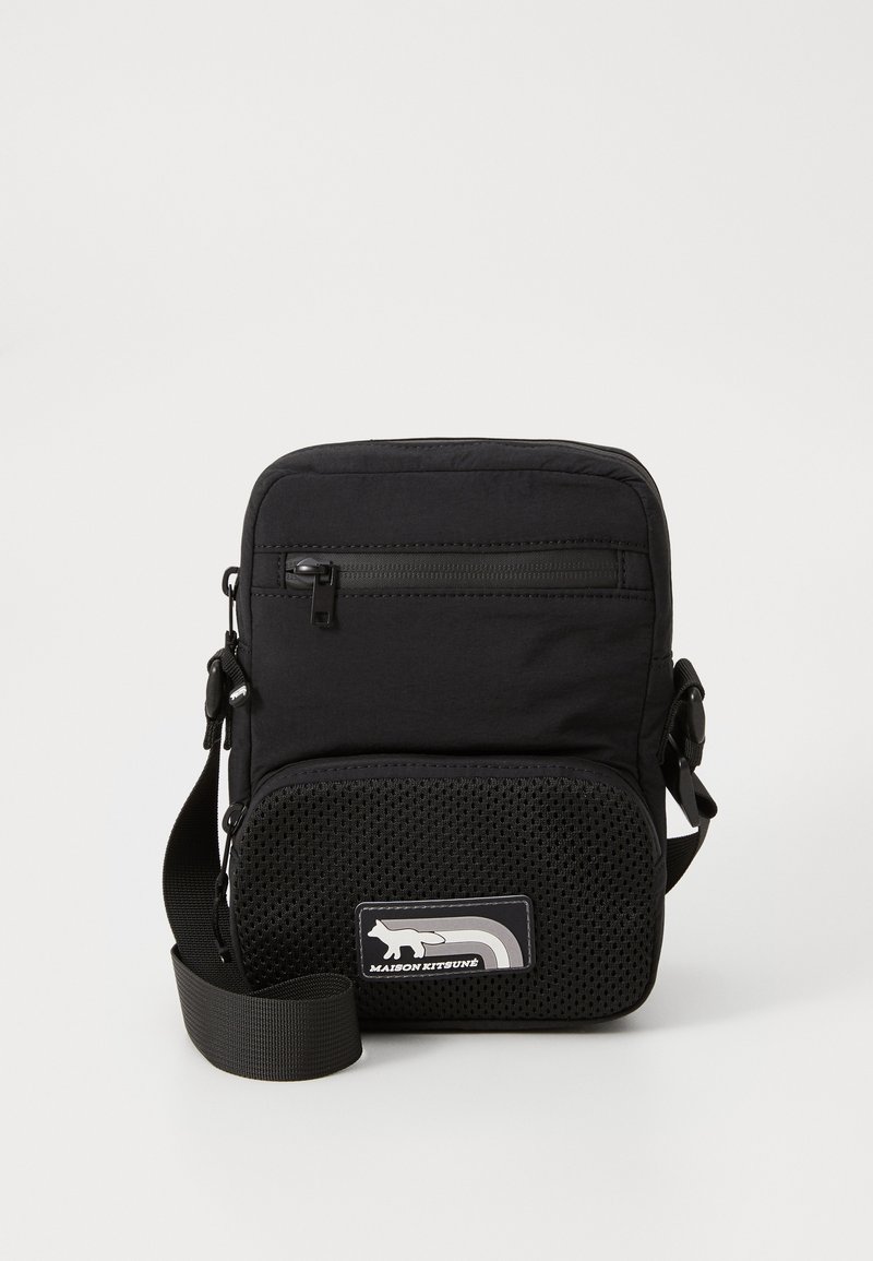 Sac bandoulière noir avec une partie supérieure en tissu doux, une section inférieure en maille texturée, une poche frontale zippée et une sangle réglable. Présente un patch logo.
