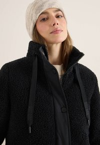 Czarna polarowa kurtka o teksturowanej powierzchni, z wysokim kołnierzem i regulowanymi czarnymi sznurkami. W połączeniu z jasnoszarym dzianinowym beanie.