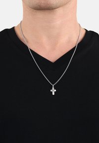 Silberne Kettenhalskette mit einem kleinen Kreuzanhänger und strahlenförmigen Details, getragen über einem schwarzen V-Ausschnitt-Shirt.