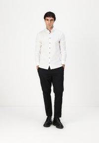 Bijela košulja s dugim rukavima i tamnim gumbima, slim fit dizajn; uparena s crnim hlačama i crnim cipelama.