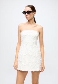 Strapless white mini dress with floral texture, layered embellishments, and a fitted silhouette; paired with black angular sunglasses.