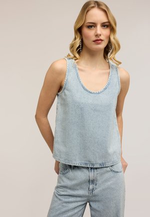 Giovane donna con capelli biondi ondulati, indossa un top di jeans azzurro chiaro senza maniche e jeans abbinati con dettagli in strass, posa davanti a uno sfondo beige.