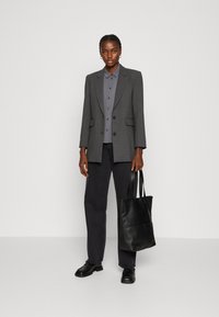 Grijze gestructureerde blazer met twee knopen, gecombineerd met een grijs overhemd en donkere jeans. Zwarte schoudertas in de hand, zwarte schoenen.