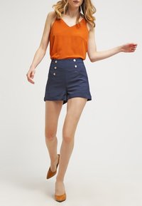 Pomarańczowy top bez rękawów z dekoltem w serek w połączeniu z granatowymi szortami z wysokim stanem, ozdobionymi srebrnymi guzikami i podwiniętymi nogawkami. Brązowe buty na obcasie.