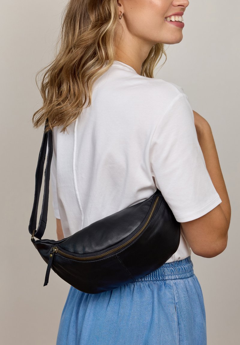 Black leather fanny pack with a curved shape, gold zipper hardware, and a flat strap, worn over a white shirt, paired with blue pants.