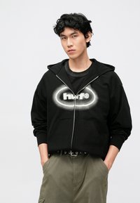 Čierna mikina na zips s bielym sprejom logom "huf®" na hrudi. Má priestrannú kapucňu a klokanie vrecko, v kombinácii s olivovozelenými nohavicami.