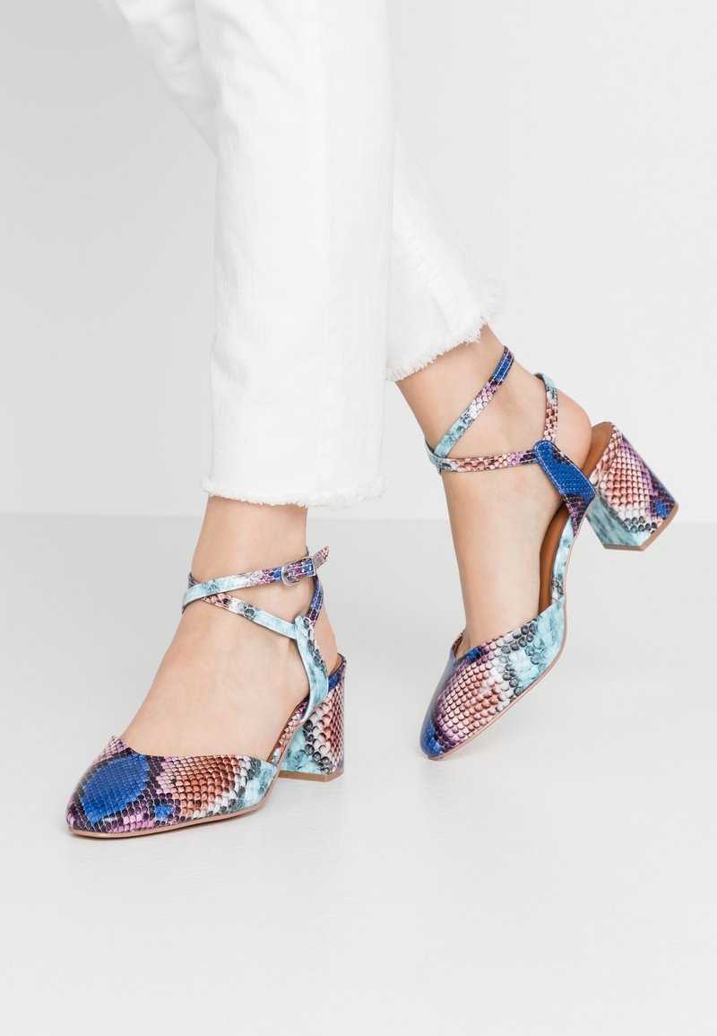 Pieds chaussés de chaussures à talons carrés à motif de peau de serpent multicolore avec des brides à la cheville, associées à un pantalon blanc à ourlet effiloché.