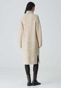 Beiges Strickkleid aus Wollmischung mit hohem Kragen, seitlichen Schlitzungen und lockerer Passform, das bis zur Hälfte der Waden reicht. Darunter werden schwarze Stiefeletten getragen.