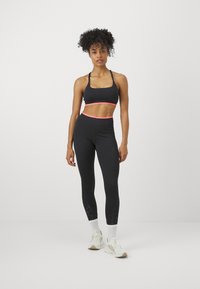 Sujetador deportivo negro con detalles en rosa y leggins negros de tiro alto que presentan un panel inferior texturizado. El modelo lleva zapatillas blancas y calcetines.