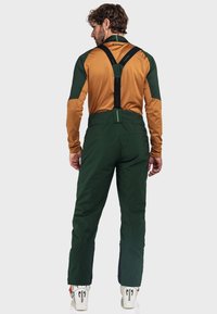 Mann in grünen Skihosen mit schwarzen Trägern, einem braun-grünen Langarmshirt und weißen Skischuhe, steht mit dem Rücken zur Kamera.