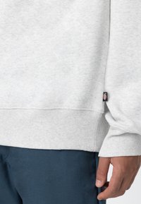 Grey sweatshirt with ribbed cuffs and hem, featuring a textured fabric and a small logo tag on the side. Worn with navy blue pants.
