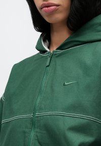Sweat à capuche zippé vert en tissu doux, avec des coutures blanches, un petit logo Nike sur la poitrine et une capuche doublée.