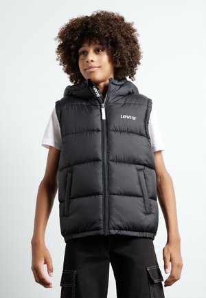 Chaleco acolchado negro con capucha, con cremallera frontal, secciones cosidas y logo en el lado izquierdo del pecho; usado sobre una camiseta blanca.