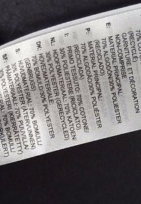 Etykieta pielęgnacyjna ubrania, pokazująca skład tkaniny w kilku językach, podkreślająca 70% bawełny z recyklingu oraz 30% poliestru.