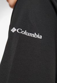 Tissu noir avec logo Columbia blanc imprimé sur la partie supérieure de la manche d'un vêtement, montrant partiellement le cou et la mâchoire d'une personne.