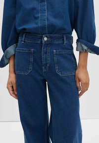 Personne portant un jean large bleu avec de grandes poches avant en patch et une chemise en denim bleu rentrée dans le pantalon, manches retroussées.