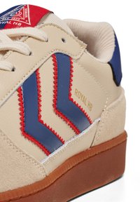 Sneaker beige con accenti a chevron blu e rossi, tomaia in pelle testurizzata, lacci chiari e suola in gomma marrone. "ROYAL HB" stampato in oro.