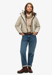 Beige cropped jacket met zwarte accenten, gebreide crèmekleurige trui, blauwe spijkerbroek en bruine veterschoenen. Model staat met handen in de zakken.