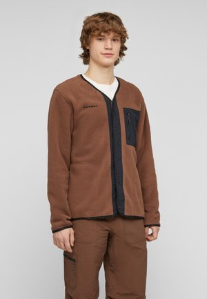 Jeune homme aux cheveux bouclés portant une veste polaire marron avec une bordure noire et un pantalon cargo marron assorti sur un fond clair uni.