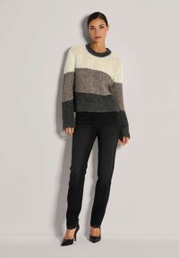 Frau mit dunklen Haaren, die einen gestrickten Pullover in Weiß, Grau und Schwarz mit Streifen, schwarze, eng anliegende Jeans mit Spitze und schwarze, hochhackige Schuhe trägt und steht.