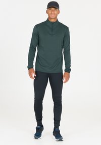 Donkergroen lange mouw quarter-zip top, aangesloten zwarte leggings en marineblauwe sportschoenen. Gladde textuur met subtiel ruitpatroon op de top.