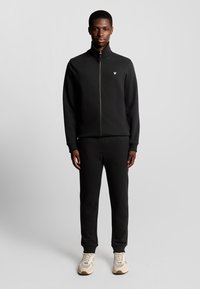 Schwarze Zip-up-Sweatjacke mit Stehkragen, kombiniert mit passenden Hosen. Das Outfit verfügt über ein dezentes Logo auf der Brust und beigen Turnschuhen.