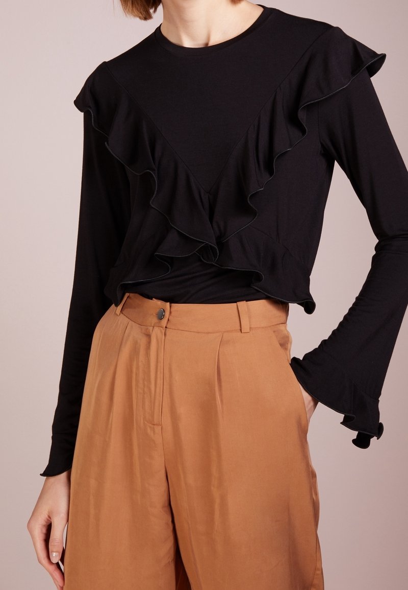 Svart långärmad topp med volangdetaljer vid halsringning och nederkant. Matchad med högmidjade, ljusbruna vida byxor.
