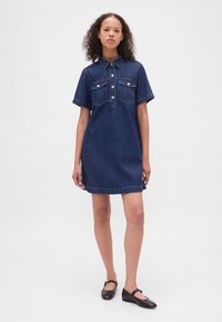 Jeansskjortklänning i mörkblått med korta ärmar, knäppning framtill, två bröstfickor och rak skärning. Bärs med svarta platta skor.