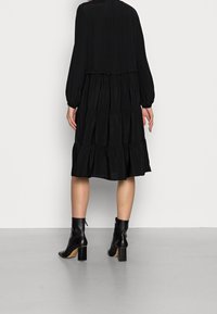 Svart lager-på-lager klänning med långa puffärmar och rund halsringning, med en flödande design och en slät textur. Kombinerad med klackade ankelstövlar.