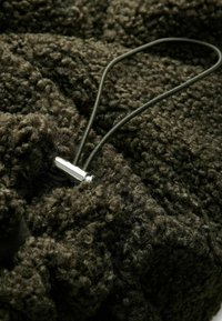 Dunkelgrüner, strukturierter Stoff mit einer samtigen, lockigen Oberfläche. Verfügt über einen silberfarbenen Metallverschluss und ein elastisches Band zur Anpassung.