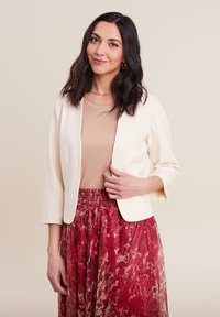 Crème kleurige blazer met driekwart mouwen, gecombineerd met een beige top en een vloeiende, multi-kleurige bloemenrok met een geplooid ontwerp.