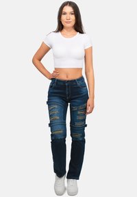 Jeans in denim blu scuro con strappi, slim fit, cuciture a contrasto e dettagli ricamati sulla coscia; abbinati a un top corto bianco.