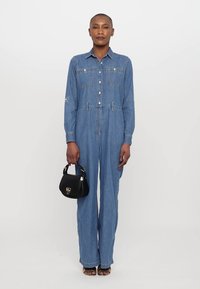 Une combinaison en denim bleu clair avec des manches longues, un devant à boutons, deux poches poitrine et un design à jambes larges. Le modèle tient un petit sac à main noir.