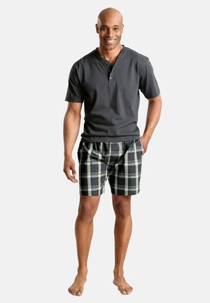 Smiling man standing barefoot wearing a dark short-sleeve henley shirt and black plaid shorts with green and white stripes.