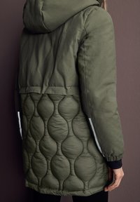 Veste rembourrée vert olive avec une section inférieure matelassée, une capuche et des accents réfléchissants sur les manches. Texture lisse avec un design superposé.