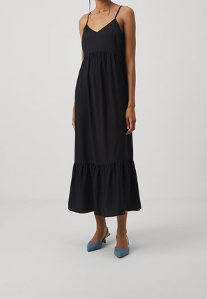 Kobieta ubrana w czarną, bezrękawną sukienkę midi z falbaną u dołu i niebieskie szpilki typu slingback z ostrym noskiem na jednolitym tle.