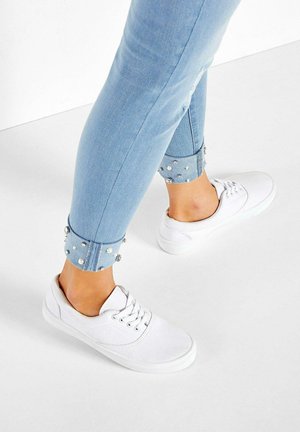 Weiße Leinenschuhe kombiniert mit hellblauen Skinny-Jeans, die an den Säumen umgeschlagen und mit silbernen Nieten verziert sind. Vor einem weißen Hintergrund platziert.