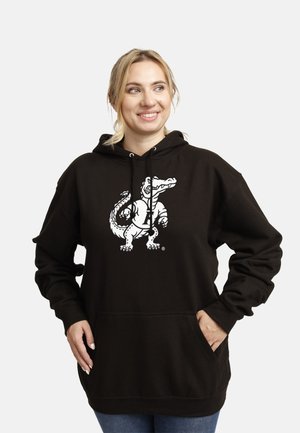 Smilende kvinde iført en sort hættetrøje med en hvid tegneseriealligators logo, hænderne i lommerne, kigger til siden mod en ensfarvet baggrund.